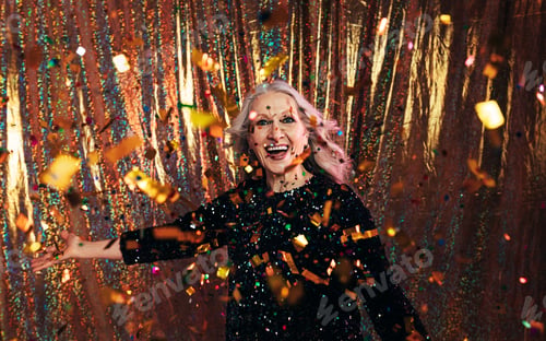Preview: Stylish senior woman in black dress throwing confetti against a golden backdrop
