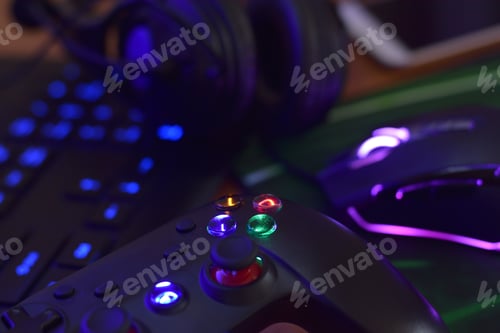 Preview: Modern gamepad and gaming mouse lies with keyboard and headphones on table in dark playroom scene