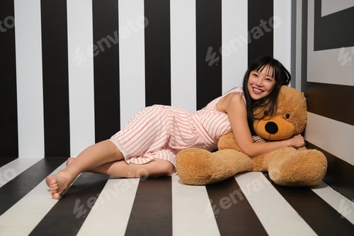 Preview: Young asian woman hug and lay over a teddy bear on the striped room floor.