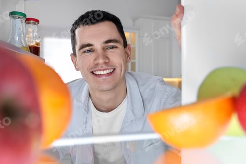 Preview: Happy man near refrigerator in kitchen, view from inside
