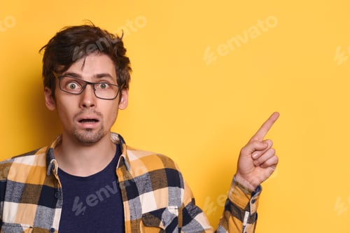 Vista previa: Joven y divertido chico nerd señalando el espacio libre para copiar texto en la pared amarilla del estudio