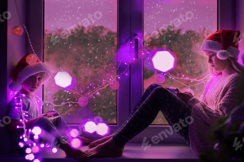 Preview: Children in Santa hat sitting on window sill and using moble phones with christmas lights background