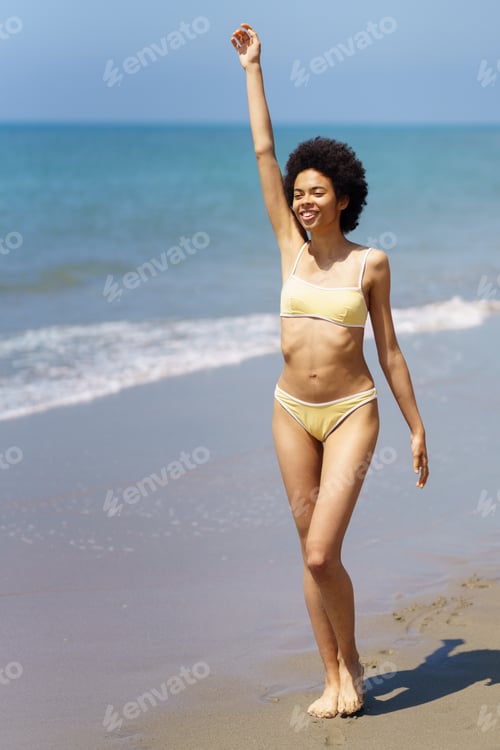 Preview: Happy ethnic woman with raised arm strolling on wet sandy seashore