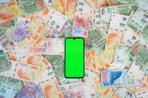 Preview: Mobile phone with green display on lots of currency notes of Argentine peso cash