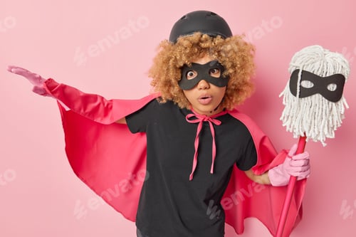 Preview: Shocked curly haired woman dressed in superhero costume with cloak wears hardhat eyemask rubber glov