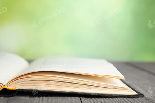 Preview: Open book on blue wooden table against blurred green background, closeup. Space for text