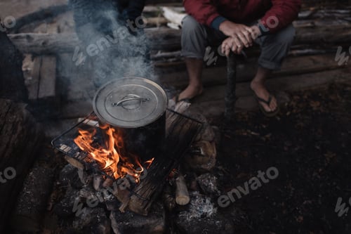 Preview: Senior man sitting on log, watching cooking pot on campfire, low section
