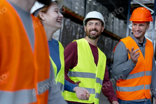 Preview: Group of smiling managers wearing hard hats and vests, walking through large warehouse, discussing