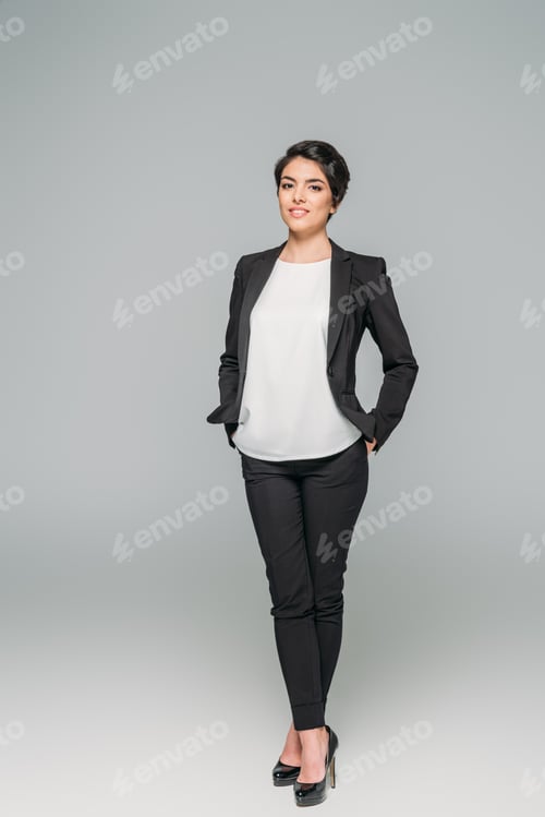 Preview: smiling mixed race businesswoman holding hands in pockets and looking at camera on grey background
