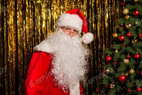 Preview: Santa claus in costume and eyeglasses looking at camera near christmas tree and golden tinsel