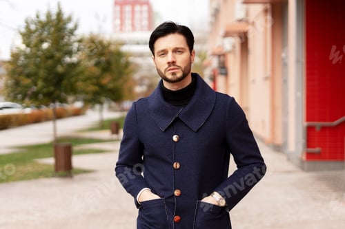 Preview: Confident young bearded man holding hands in pockets looking at camera in the city