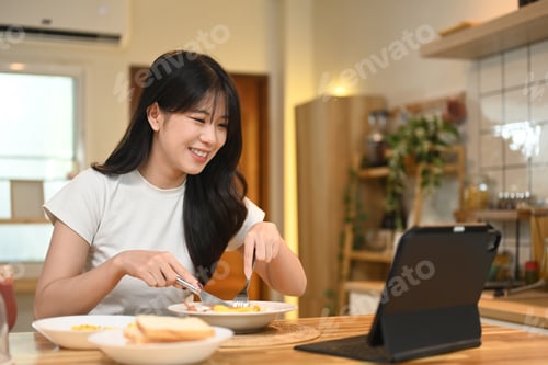 Preview: Happy young woman enjoying a meal while watching something on digital tablet at a kitchen table