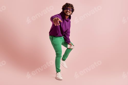 Preview: Young Indian man in headphones pointing camera and smiling against pink background