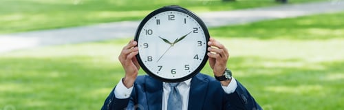 Preview: panoramic shot of businessman covering face with clock while standing in park