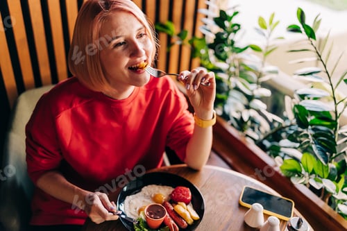 Preview: Young happy smiling woman eating breakfast in the cafe. Potatoes and sausages ketchup
