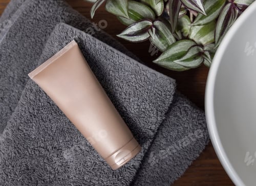 Preview: Beige cosmetic tube on grey folded towel near green plant on wood in bathroom, mockup