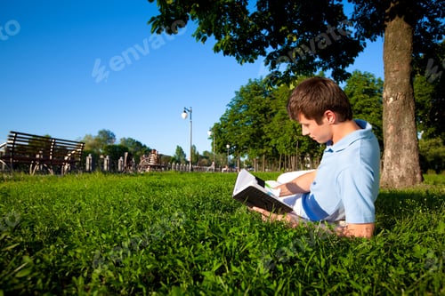 Preview: Young man lying on green grass in trees and reading book