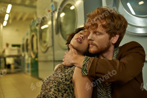 Preview: sexy young man in stylish outfit touching asian girlfriend in dress in public laundry in evening
