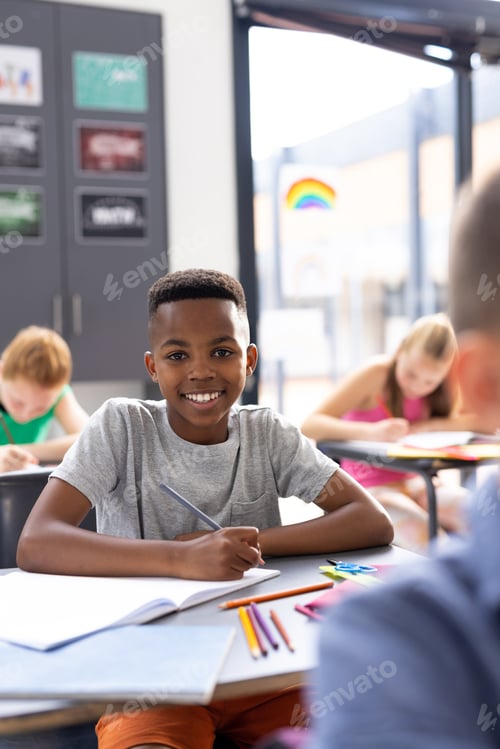 Preview: Vertical portrait of smiling african american schoolboy working at his desk in classroom, copy space
