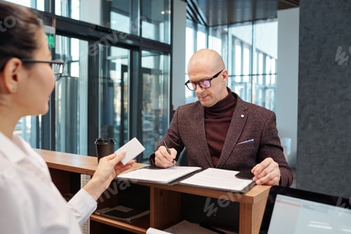 Preview: Bald confident businessman filling in form by counter in front of receptionist