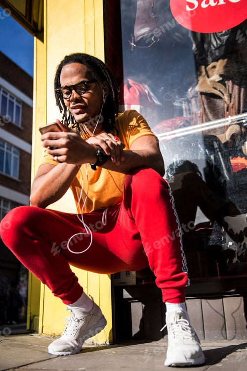 Preview: Young man crouching outdoors, wearing earphones, using smartphone