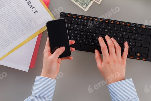 Preview: Female hands using smartphone while typing on computer keyboard.