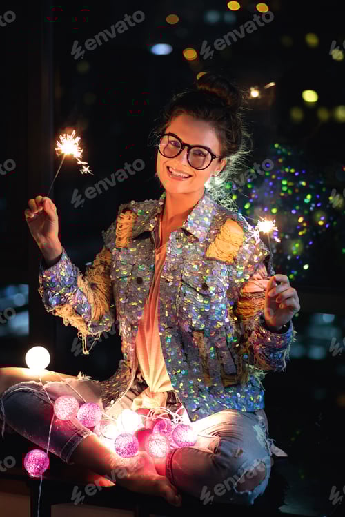 Preview: Happy woman wearing glowing jacket with sequins is holding sparklers in her hands.