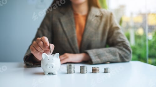 Preview: A businesswoman putting coin into piggy bank with coins stack on the table