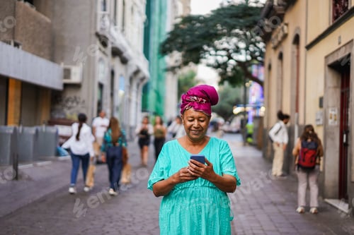 Preview: Senior african woman using mobile phone in the city while wearing traditional dress and turban