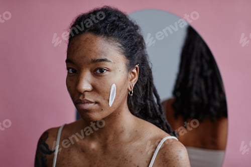 Preview: Candid young black woman with face cream smudge looking at camera by mirror