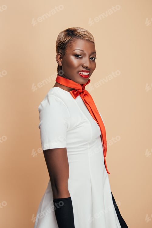 Preview: smiling african american model posing in white dress and red scarf isolated on beige