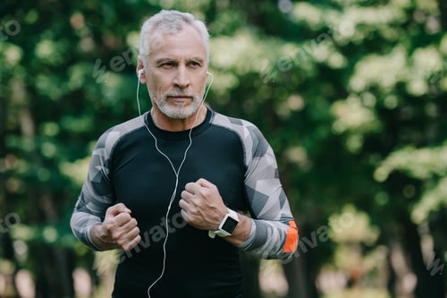 Preview: handsome mature sportsman listening music in earphones while running in park