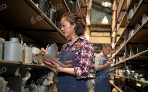 Preview: Woman in apron checking inventory on clipboard in a warehouse aisle