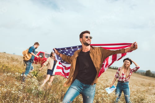 Preview: handsome young man holding usa flag and walking by field with friends during trip