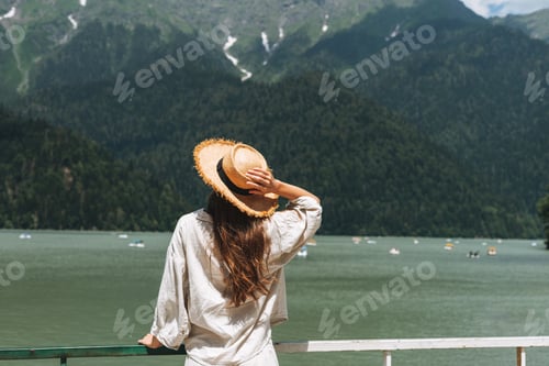 Preview: Young woman traveler with long blonde hair in straw hat looking at mountain lake