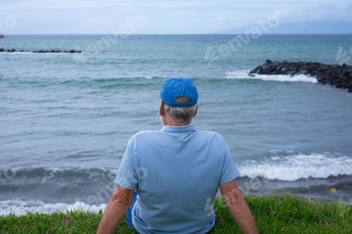 Preview: Rear view of senior man sitting on the grass at the beach in a cloudy day enjoying free time vacatio