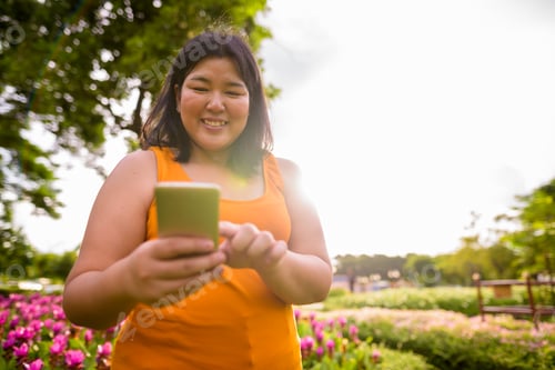 Preview: Beautiful happy overweight Asian woman using phone in park
