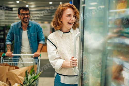 Preview: Happy couple grocery shopping, woman opening refrigerator door in supermarket,
