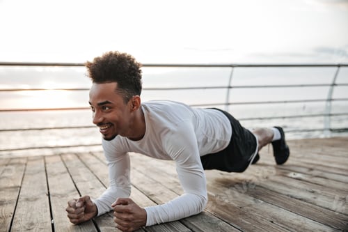 Preview: Happy excited active dark-skinned man in black shorts and white t-shirt smiles and does plank. Cool