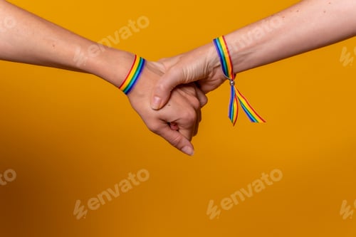 Preview: Detail of two hands of two women shaking hands with a bracelet, LGBT concept on a yellow background