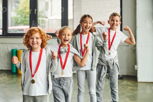 Preview: four joyful children in sportswear with medals cheering and smiling happily at camera, child sport