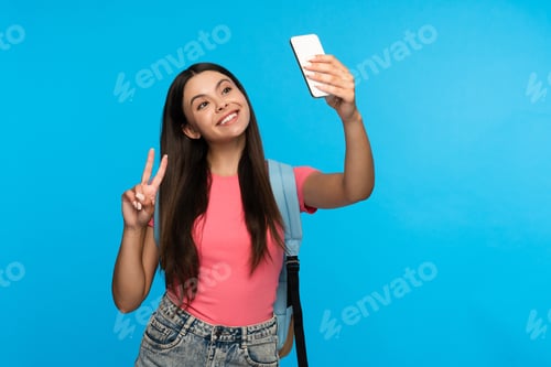 Preview: Positive girl teenager blogger takes selfie photo holding mobile phone smiling doing peace victory