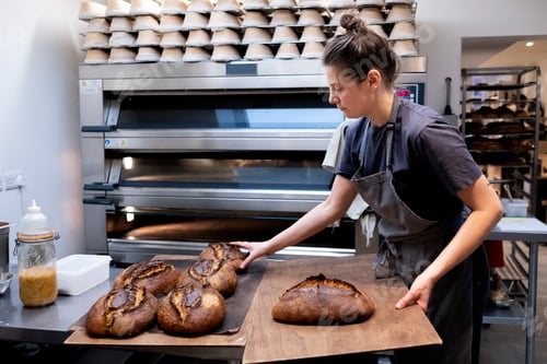 Preview: Woman wearing apron standing in an artisan bakery, placing freshly baked loaves of bread onto wooden
