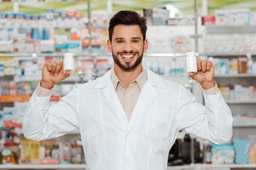 Preview: Handsome pharmacist smiling at camera and holding jars with pills
