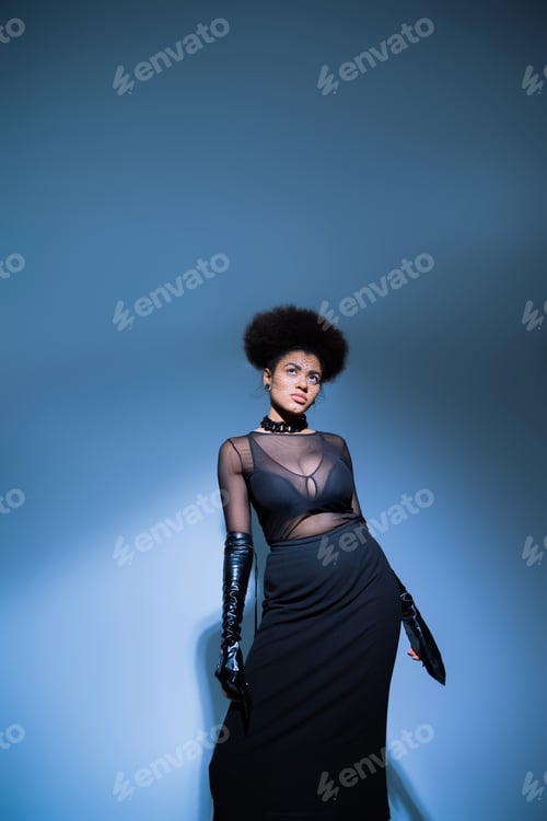 Preview: curly african american woman in transparent shirt and skirt looking up on blue