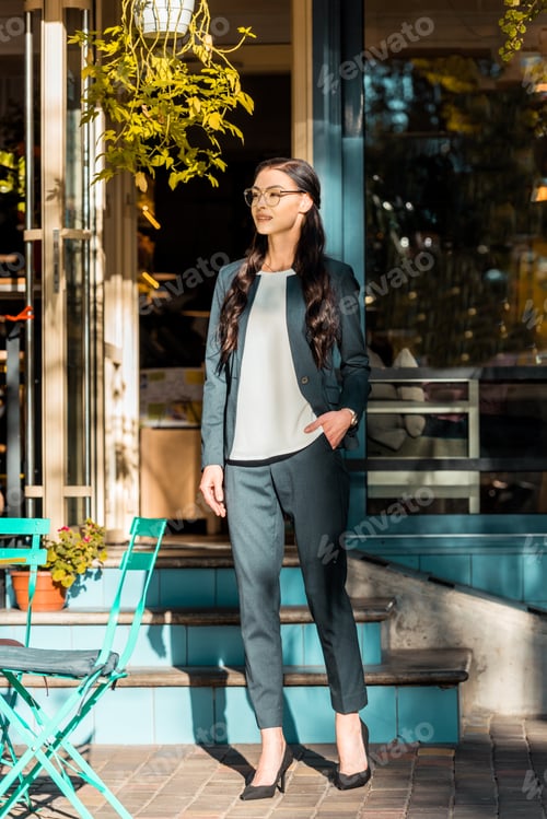 Preview: beautiful brunette businesswoman standing near stairs to cafe on street and looking away