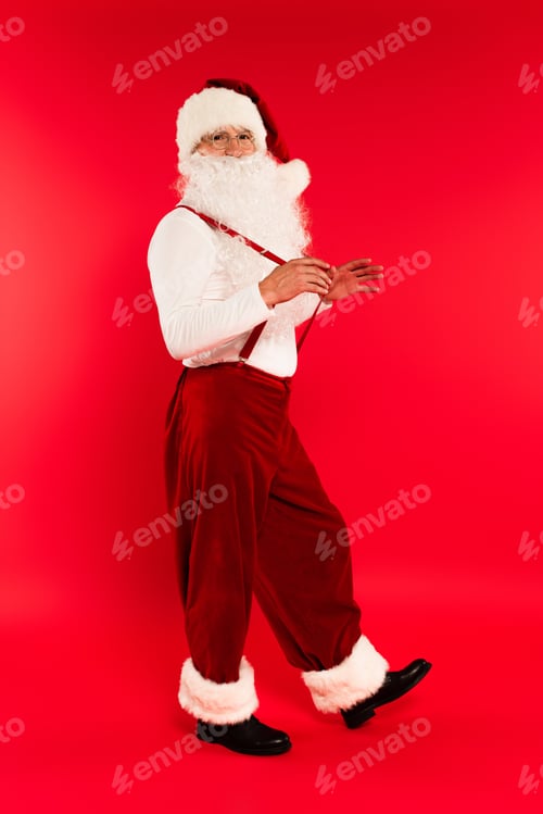Preview: Full length of santa claus in eyeglasses touching suspenders on red background