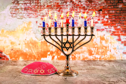 Preview: Menorah with lit candles, kippah on rustic table against an orange brick wall during a Jewish