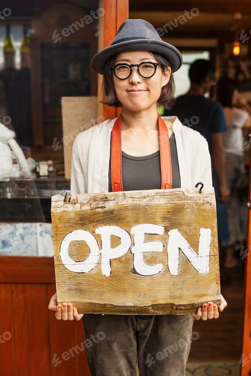 Preview: Japanese woman wearing hat and glasses standing in front of a leather shop, holding Open sign.