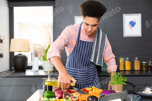 Preview: Biracial man wearing apron cooking dinner, pouring vegetables with oil in kitchen, unaltered